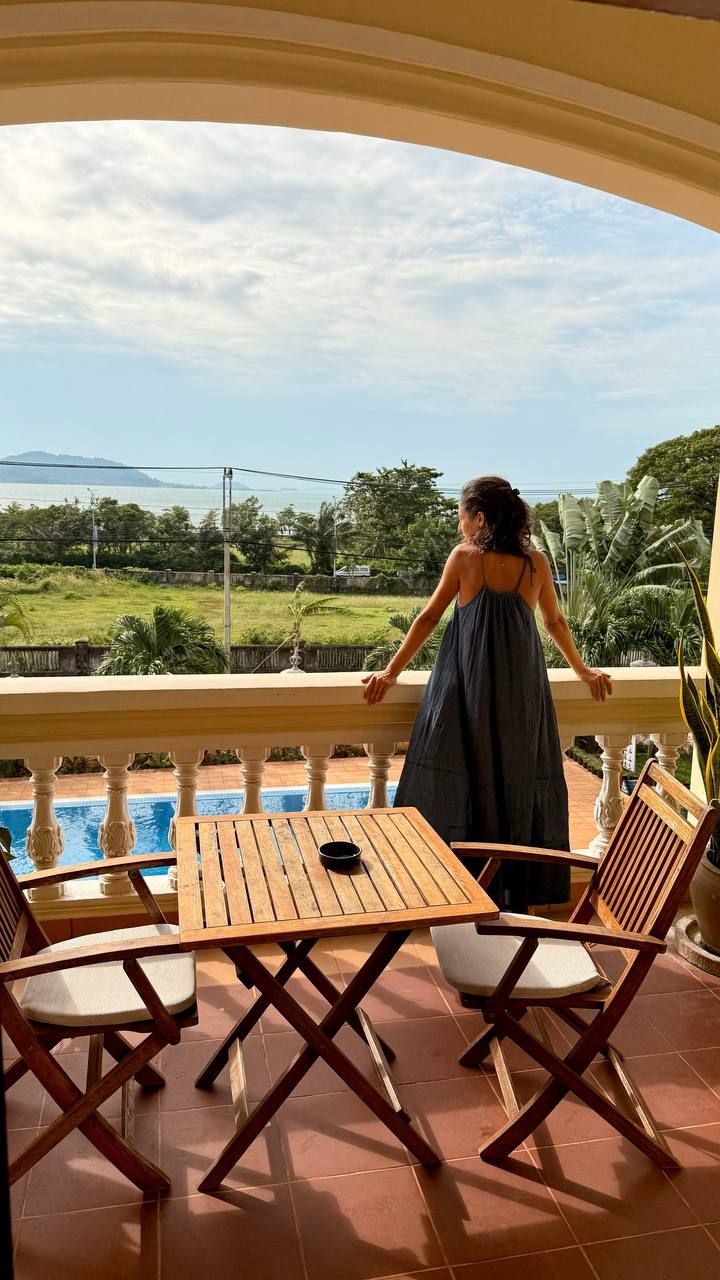 Eine Frau in einem dunklen Kleid steht auf einem Balkon mit Pool und Stühlen.