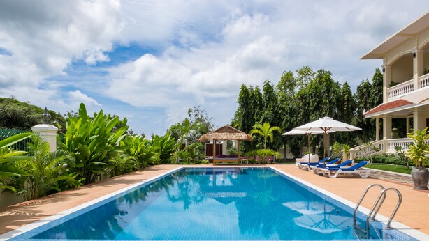 A swimming pool with lounge chairs and umbrellas, surrounded by lush greenery and a thatched-roof structure. - Riviera Hotel & Resort Kep (Kep)