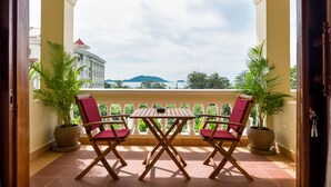 A balcony with two wooden chairs and a table, potted plants, and a view of a building and mountains. - Riviera Hotel & Resort Kep (Kep)