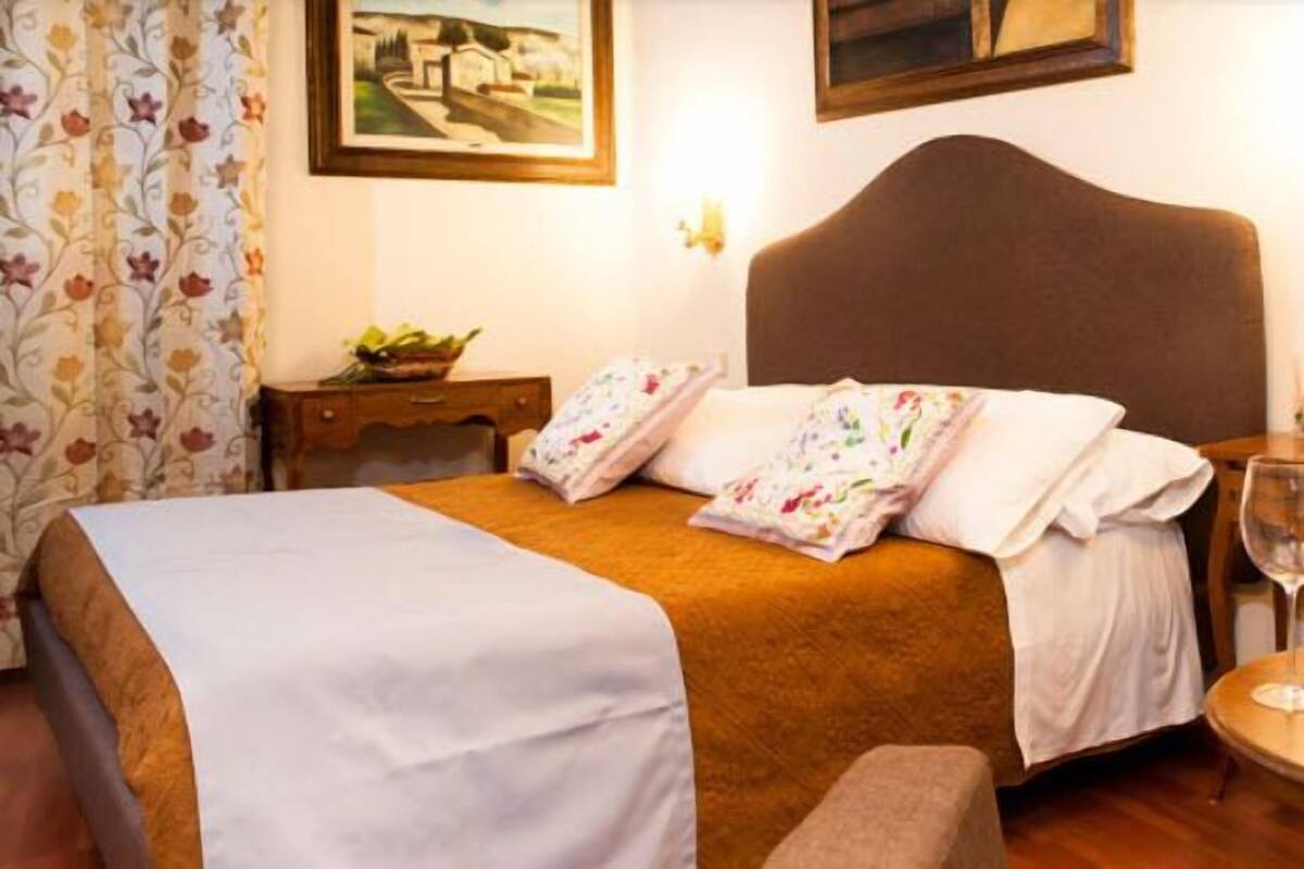 A hotel room with a bed, a bedside table with a bottle of wine and a glass, a small table with a bottle of wine, a sofa, and a painting on the wall.