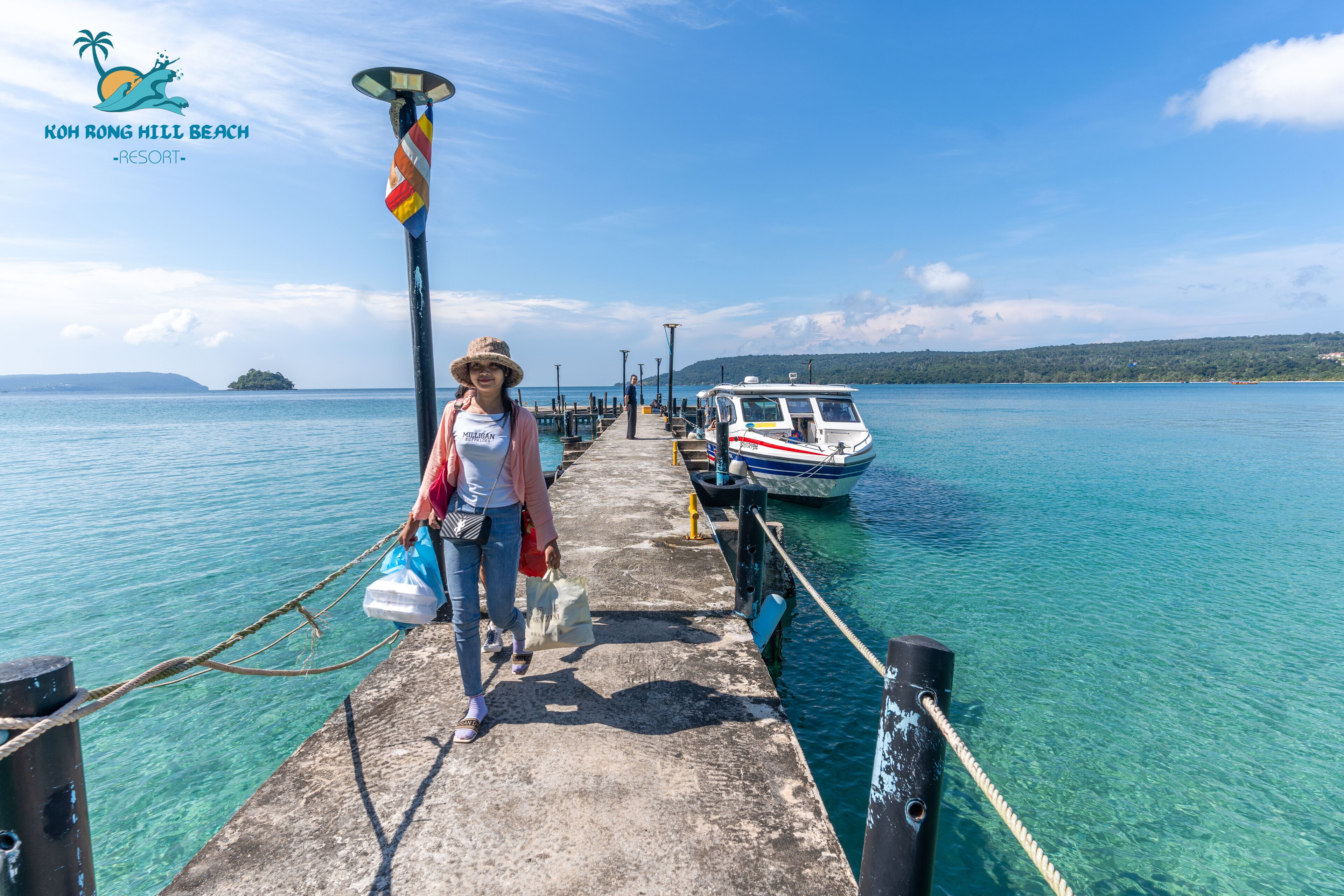 A pier at Koh Rong Hill Beach Resort with clear turquoise water, a boat docked on the right, and a person walking towards the camera.