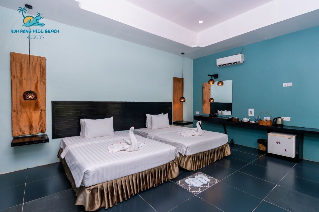 Habitación de hotel con dos camas, cabecero de madera, pared de acento color turquesa, kitchenette pequeña y ventilador de techo.
