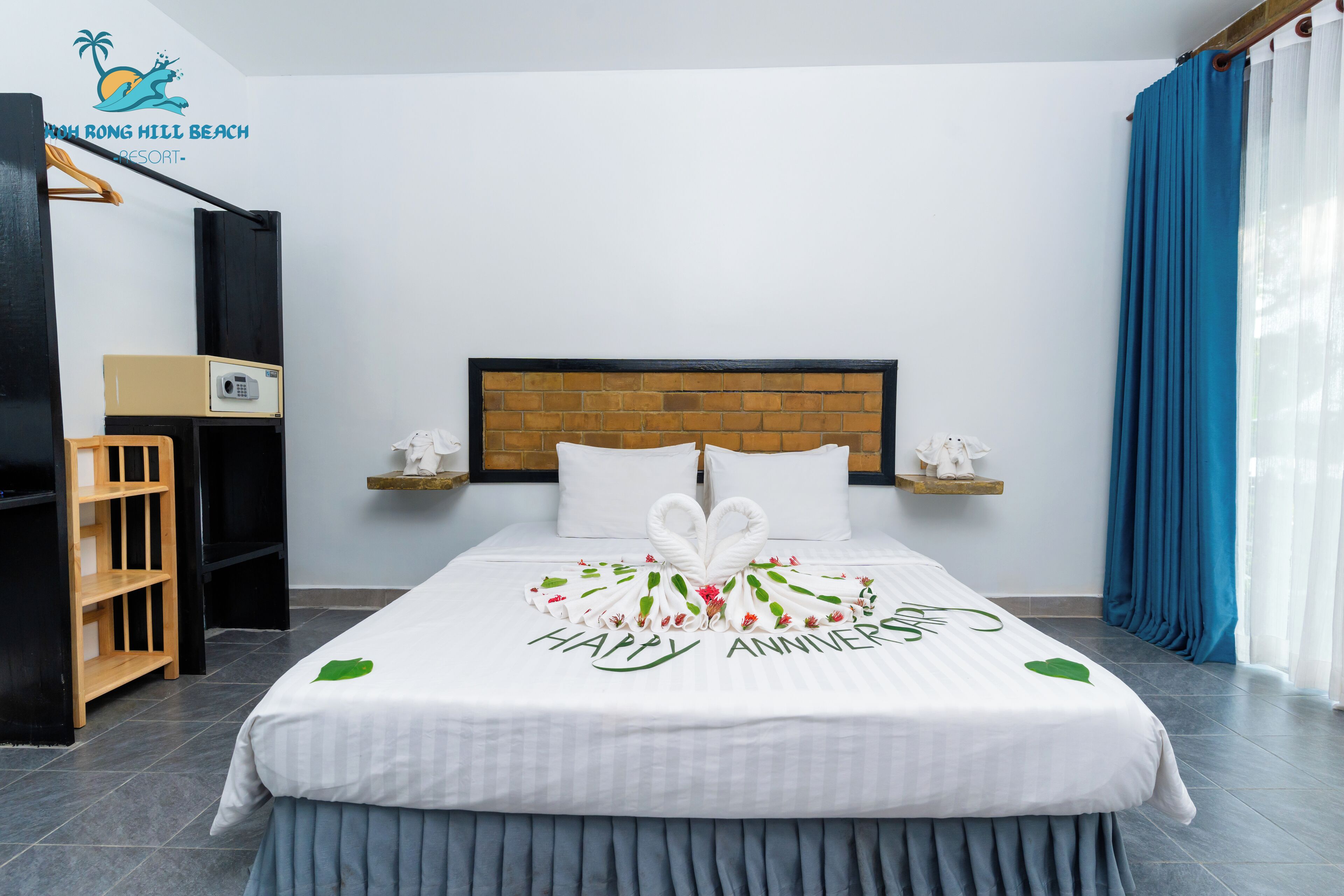 A hotel room with a bed featuring a 'Happy Anniversary' decoration, a wooden headboard, a bedside table with a phone, and a blue curtain.