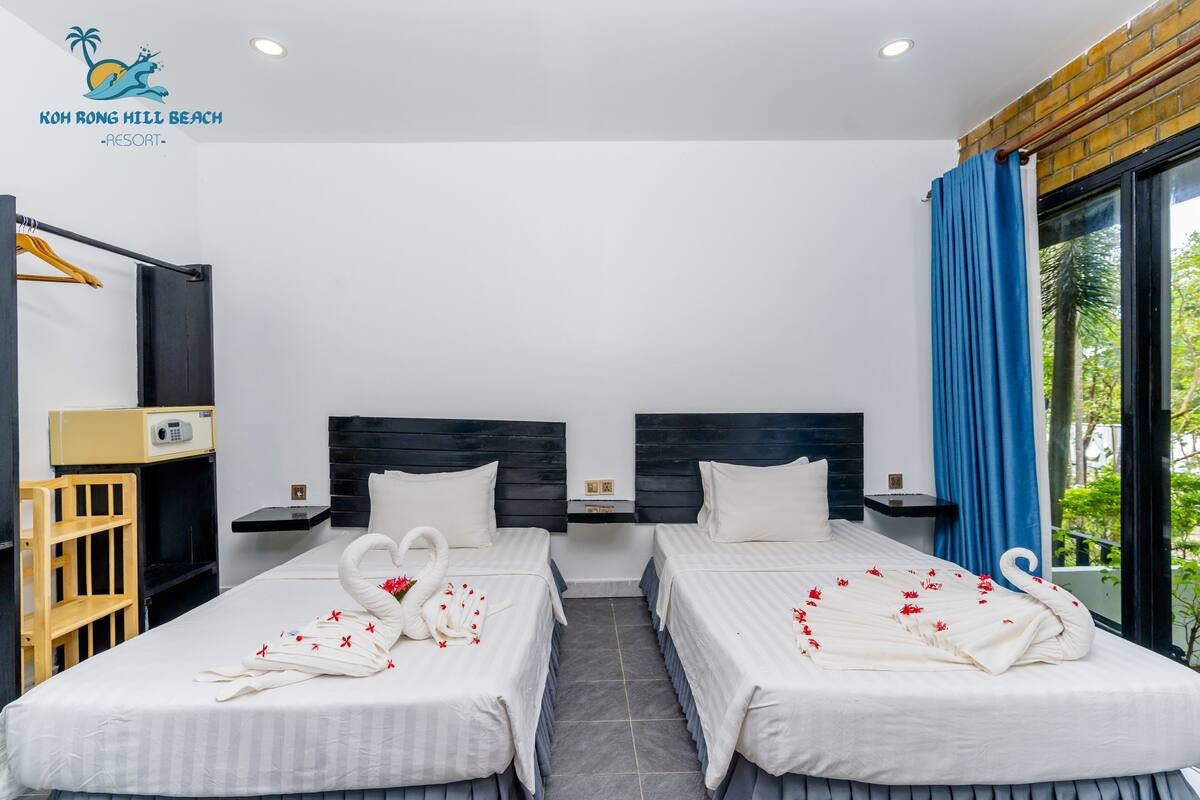 Habitación de hotel con dos camas, un escritorio pequeño, una silla, un armario y ventana con vistas a zonas verdes.