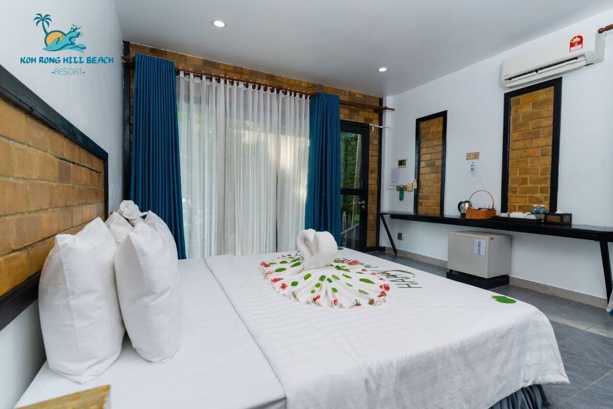 Una habitación de hotel con cama, almohadas, un escritorio, un espejo y un refrigerador pequeño.