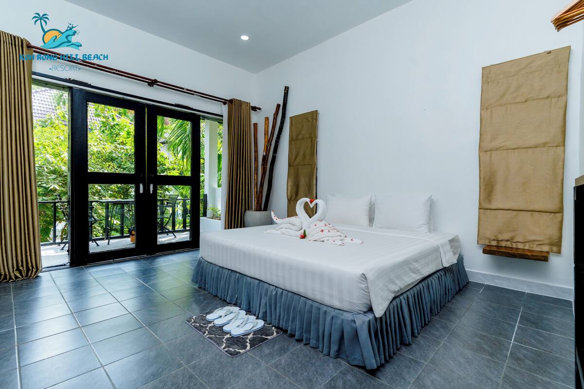 Habitación de hotel con una cama grande, vista a zonas verdes a través de una puerta corrediza de vidrio y un balcón con baranda de madera.