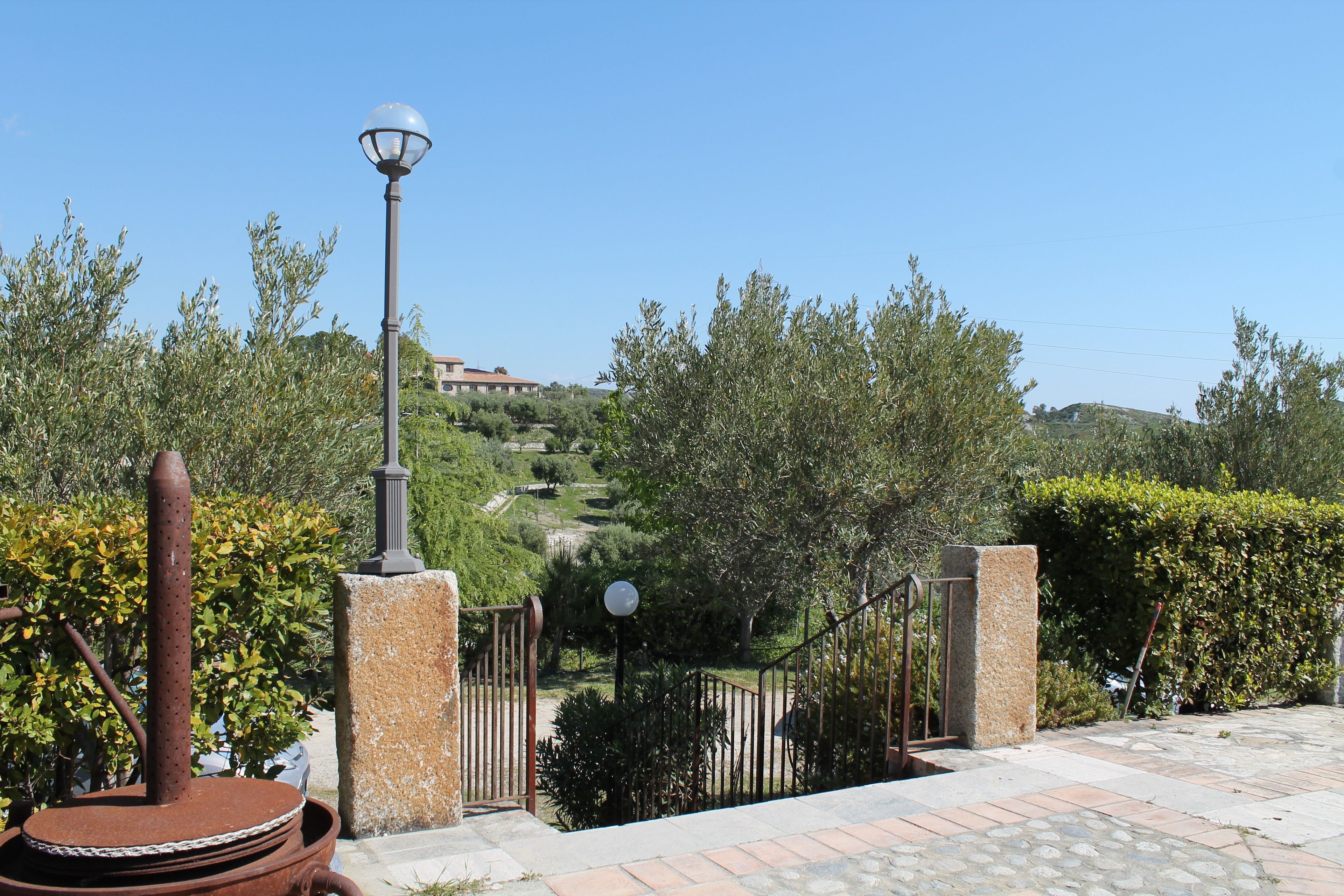 Un camino pavimentado que lleva a una entrada con puerta de rejas, un farol y una estructura metálica.