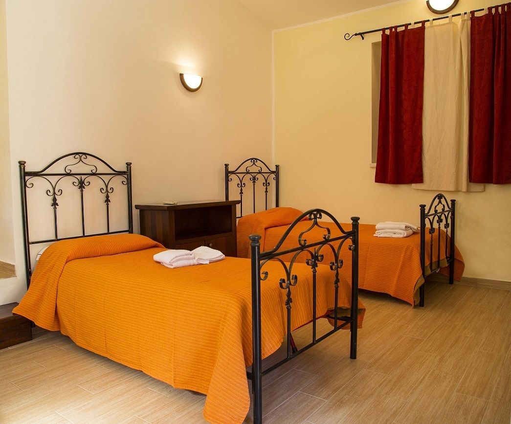 Una stanza con due letti, ognuno con copriletto arancione e cuscini bianchi, un comodino in legno e un punto luce a parete.