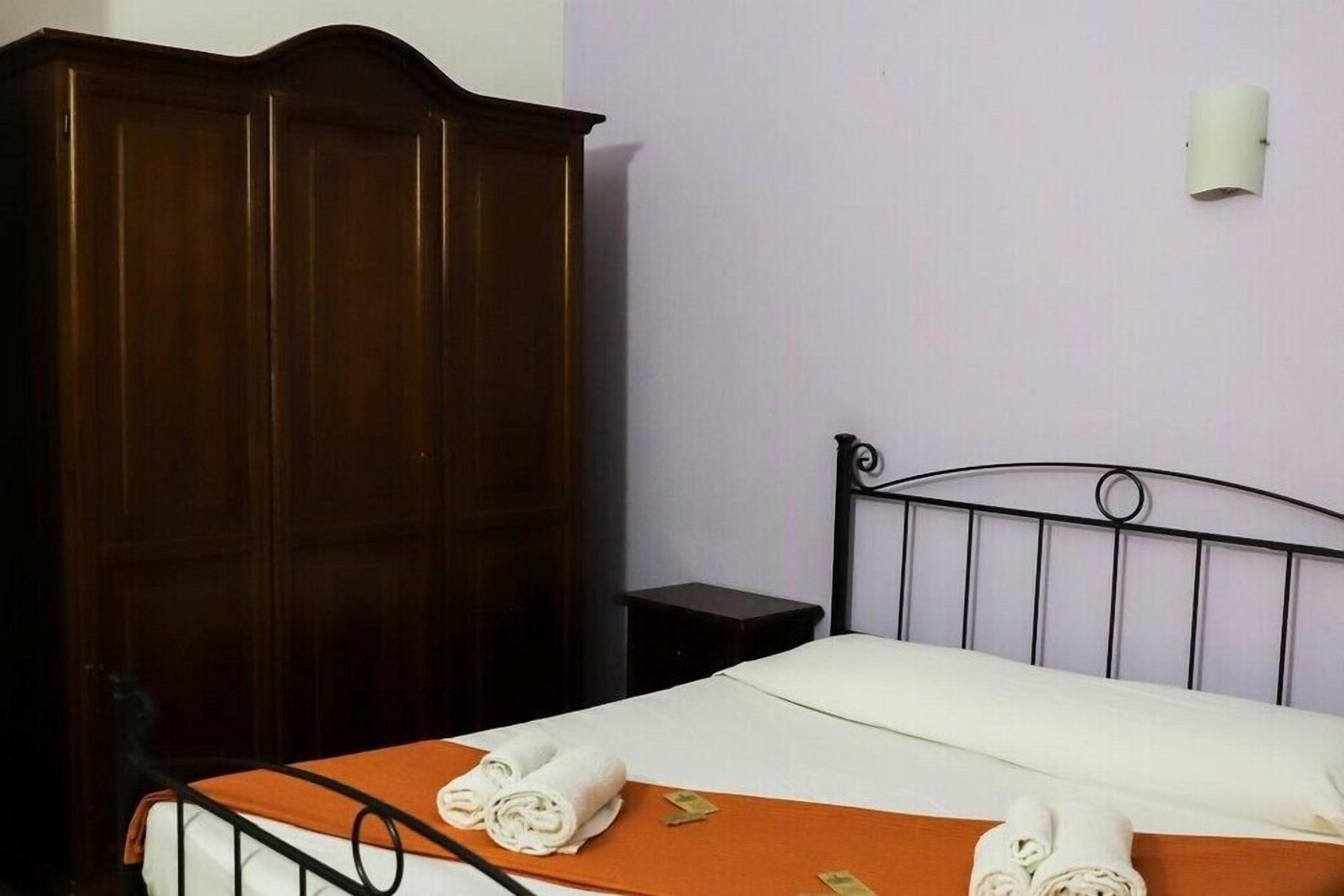 Una cama individual con estructura metálica, una colcha naranja y dos toallas enrolladas sobre la cama. A la izquierda, un armario de madera oscura y, junto a la cama, una mesita de noche negra.