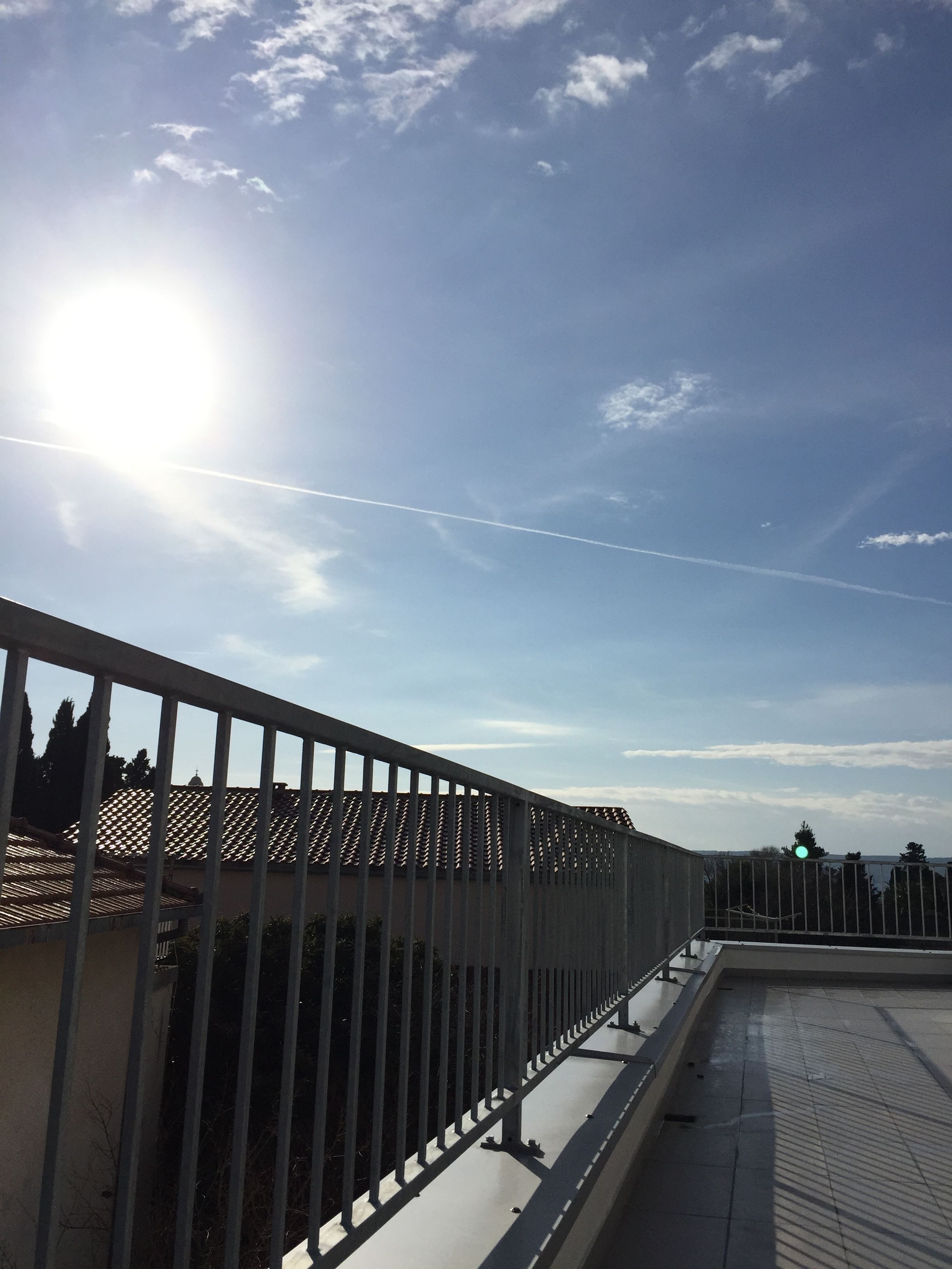 A sunny day with a clear sky, a metal fence, and a building with a tiled roof.