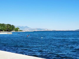 Vista costera con un puerto deportivo, mar tranquilo y un horizonte urbano lejano con montañas.