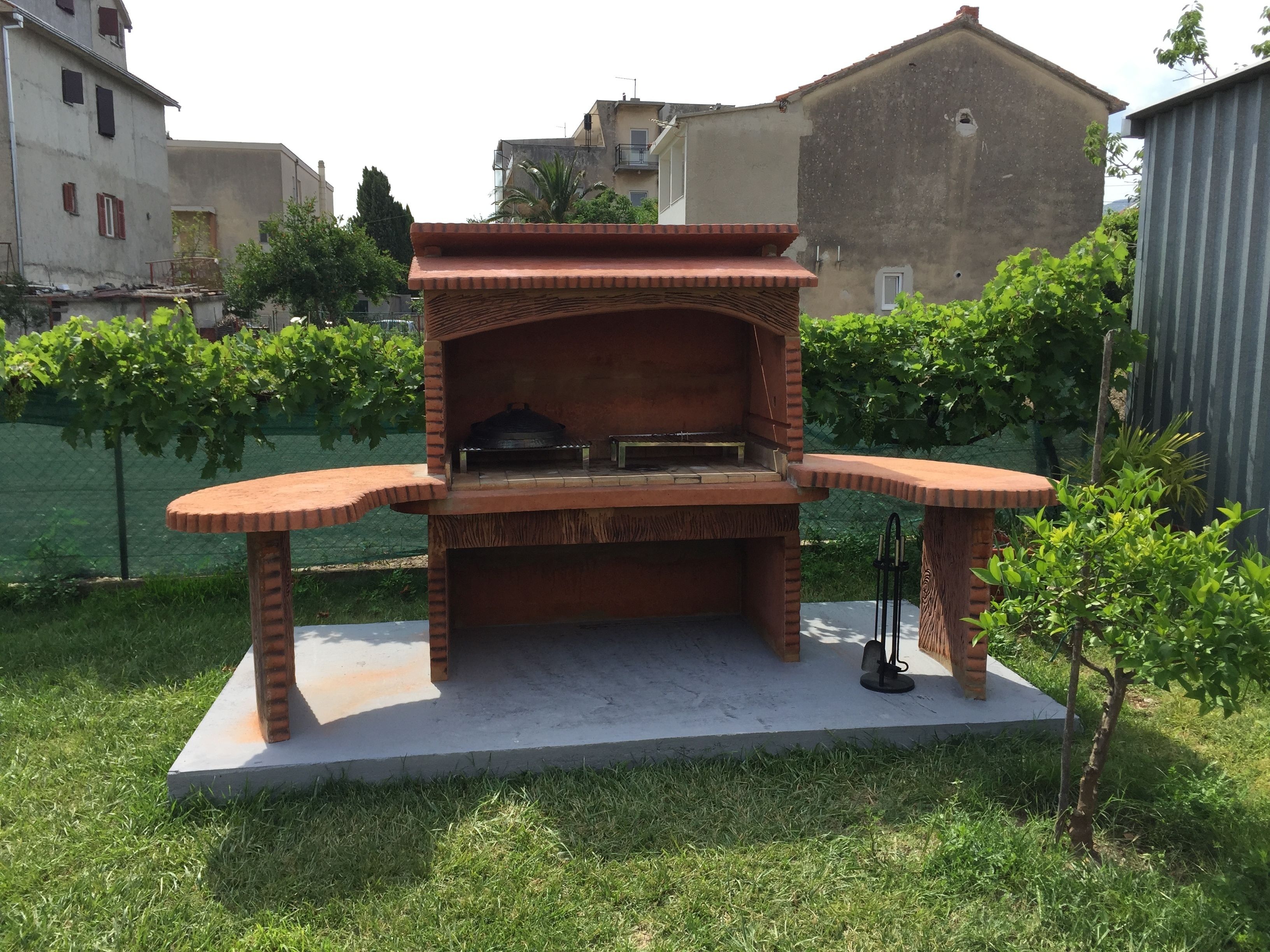 Ein gemauerter Grill mit Schornstein, auf einem Betonfundament stehend, umgeben von Gras und Bäumen.