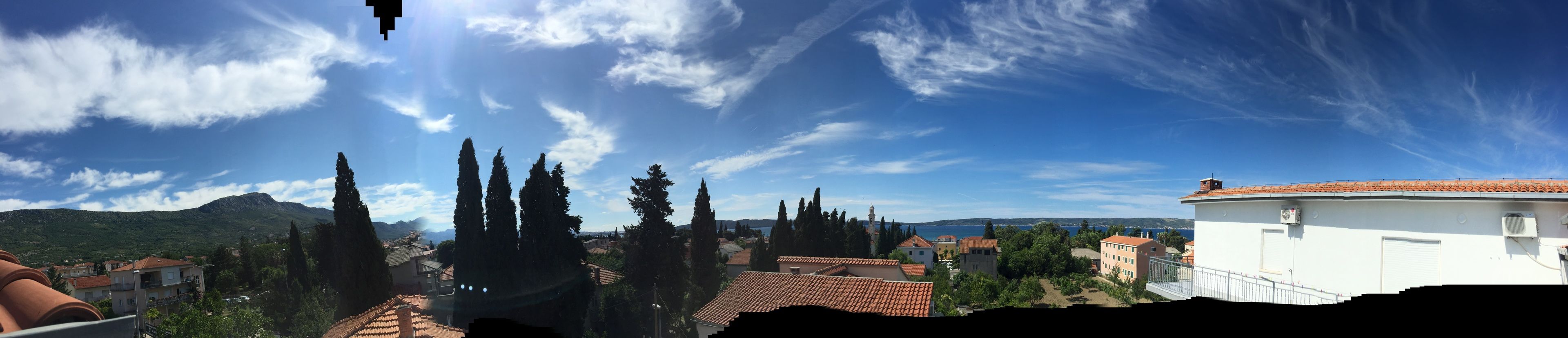 A view of a coastal town with terracotta-roofed buildings, a white building with a red roof, and a clear blue sky with scattered clouds.