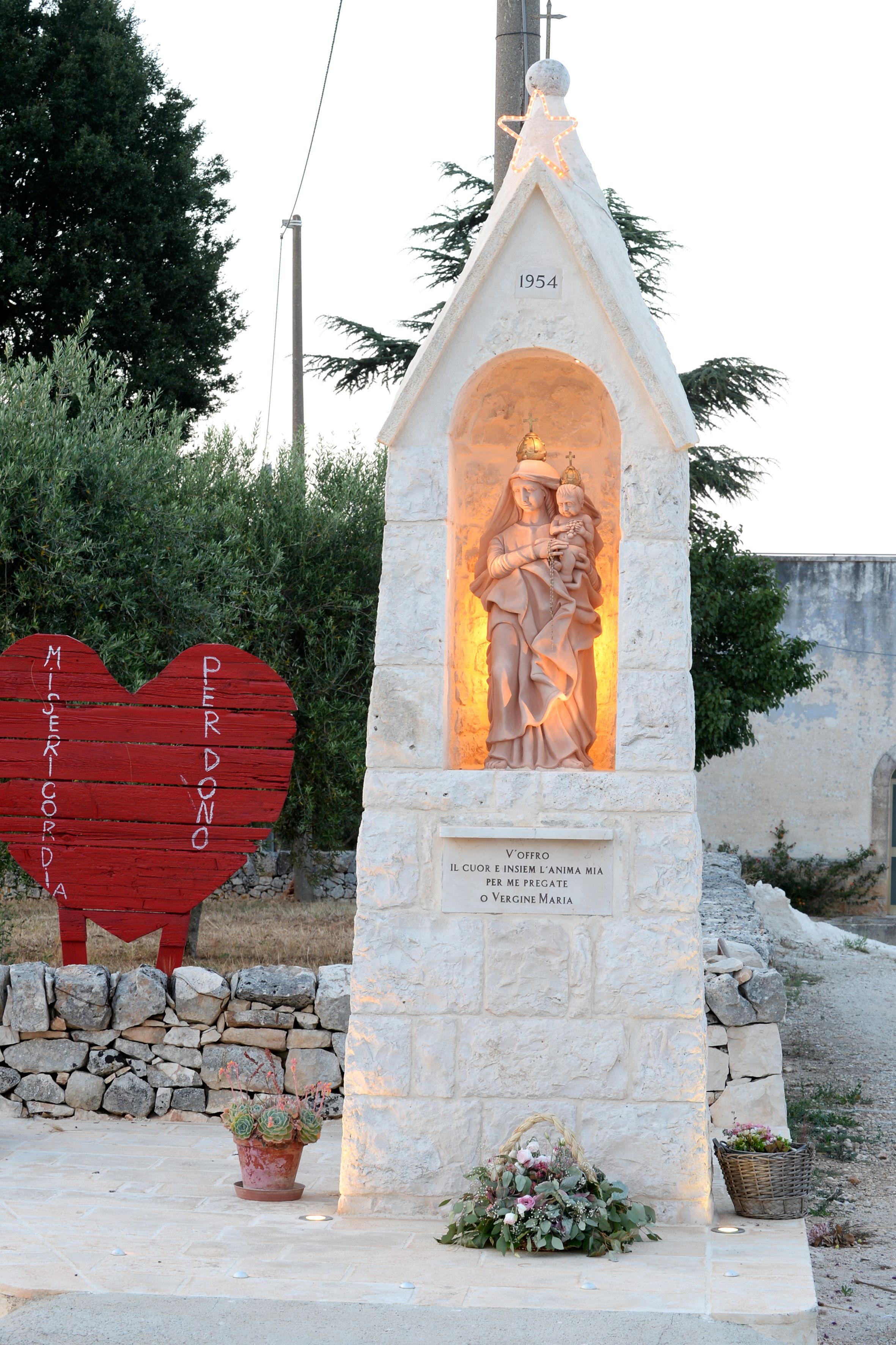 A stone monument with a statue of a religious figure, a red heart-shaped sign, and potted plants.