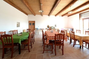 Un comedor con mesas y sillas de madera, una cocina al fondo y un techo alto de madera.