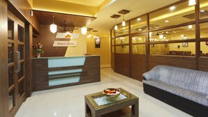 A hotel reception area with a wooden reception desk, a sofa, and a small table with a decorative bowl. - Hotel Atria (Kolhapur)