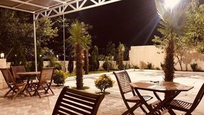 Un área de patio al aire libre con mesas y sillas de madera, rodeada de palmeras y una valla blanca.