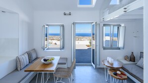 A modern, open-plan room with a view of the sea, featuring a dining area, seating, and a small table with a bowl of fruit. - Sandy & Salty Villas (Santorini)