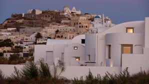 A coastal town with white buildings, some with blue domes, and a hill with more buildings. There are visible windows and doors, and some buildings have balconies. - Sandy & Salty Villas (Santorini)