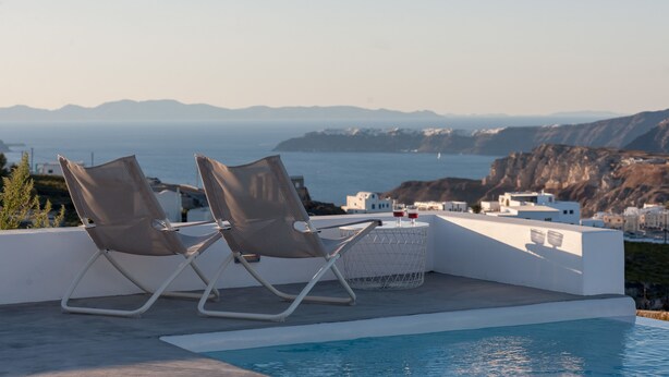 A poolside area with two lounge chairs and a small table, overlooking a coastal landscape with buildings and mountains in the distance. - Sandy & Salty Villas (Santorini)