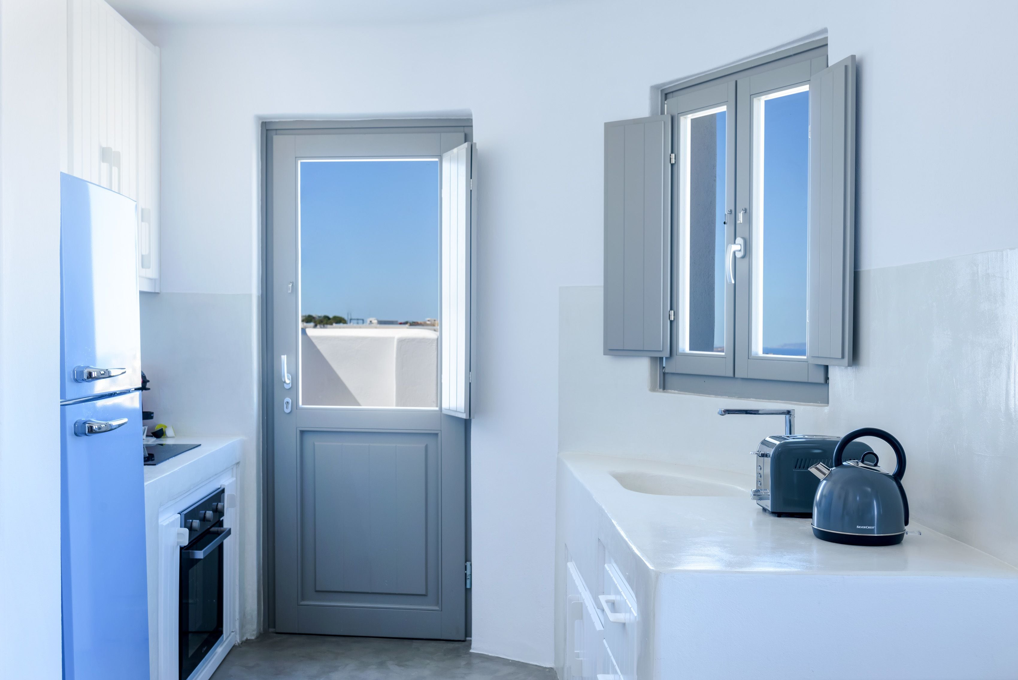 A narrow kitchen with a door leading to a balcony, a white sink, and a kettle on the counter.