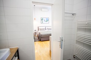 Ein schmaler Flur führt zu einem Schlafzimmer mit einem Bett, einem kleinen Tisch und einem Heizkörper.