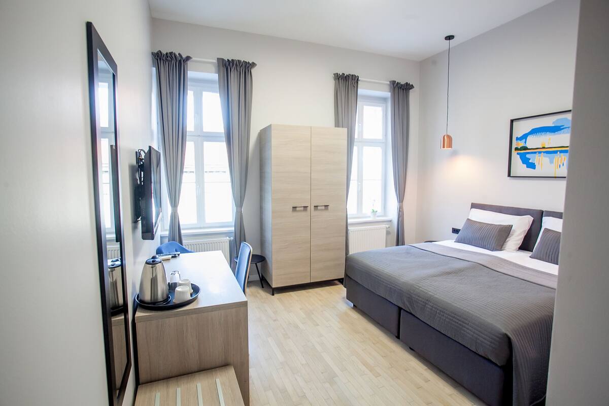A modern hotel room with a bed, a desk with a chair, a wardrobe, and a large mirror.