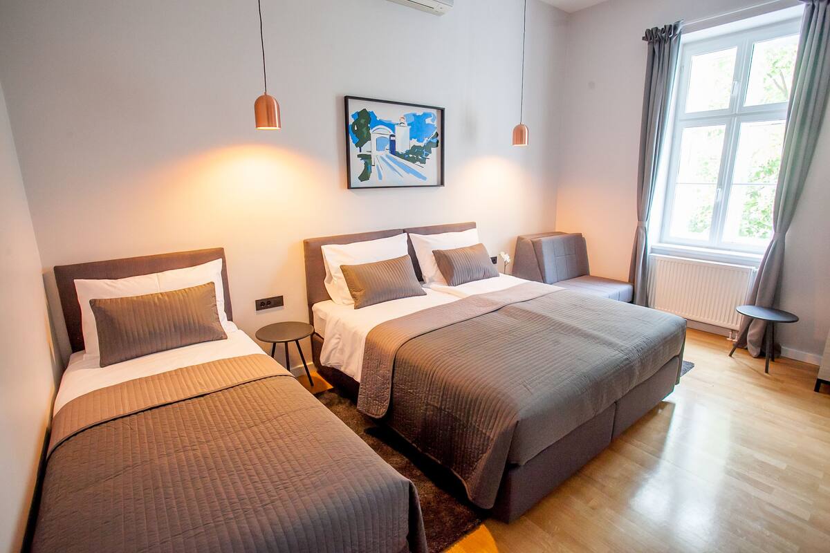 A hotel room with two beds, a small table, a chair, a window with curtains, and a painting on the wall.