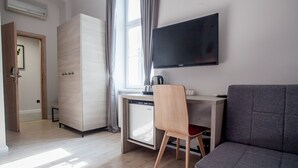 A modern hotel room with a wooden floor, a grey sofa, a wooden desk with a chair, a television, and a minibar. - Zagreb City Vibe Apartments & Rooms (Zagreb)