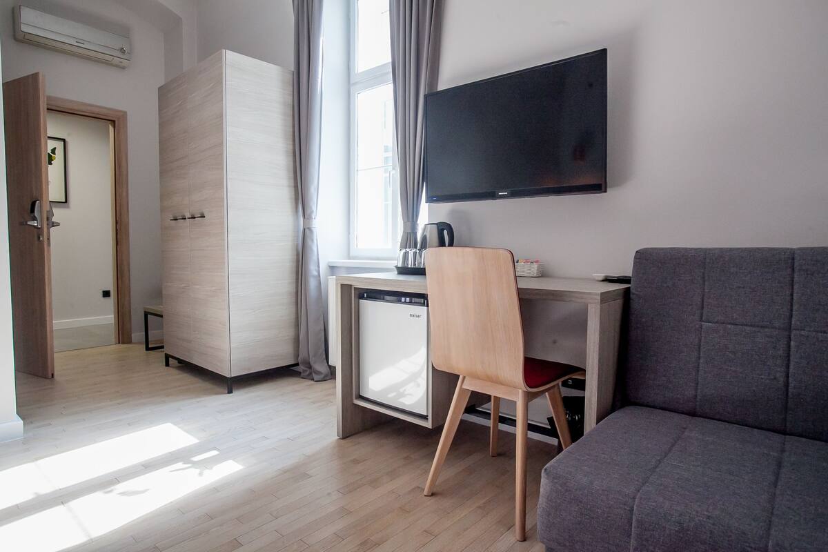 A modern hotel room with a wooden floor, a grey sofa, a wooden desk with a chair, a television, and a minibar.