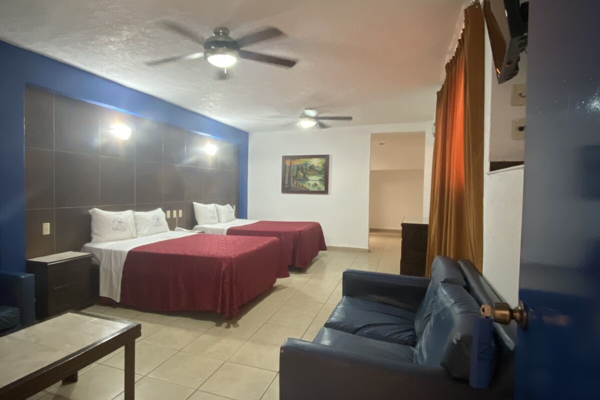 Una habitación de hotel con una cama, dos mesitas de noche, un escritorio, una silla, un sofá, un ventilador de techo y un cuadro en la pared.