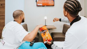 Two people sitting on a couch watching TV, with a bag of Doritos and a remote control visible. - Rotterdam Teleport Hotel (Rotterdam)