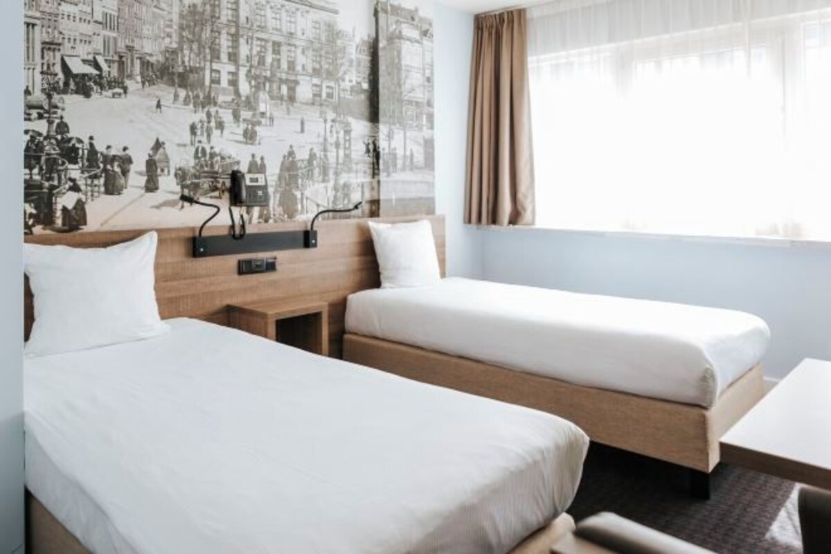 A hotel room with two beds, a wooden headboard, a nightstand, and a large window with curtains.