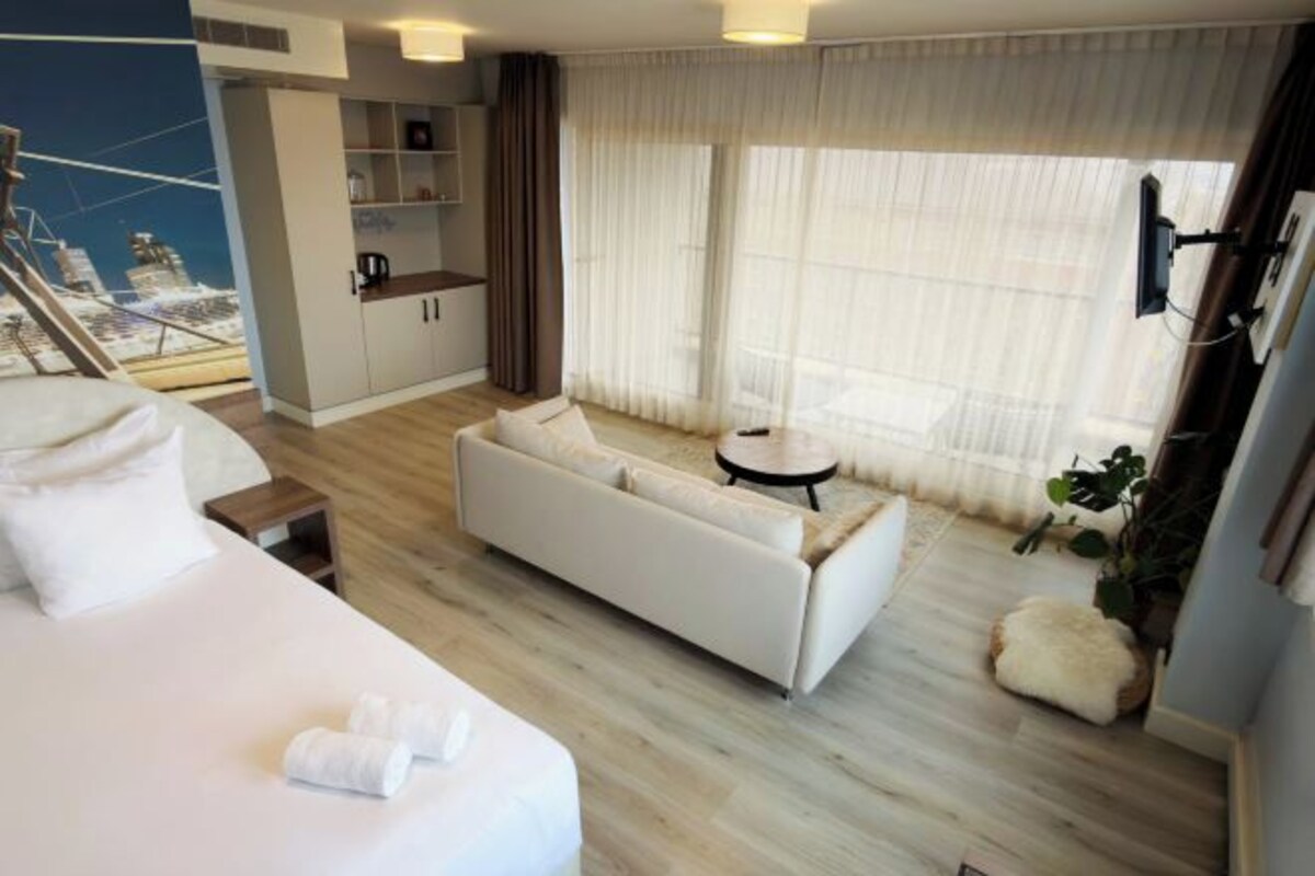 A hotel room with a bed, a sofa, a small table, a plant, and a view of a cityscape through sheer curtains.