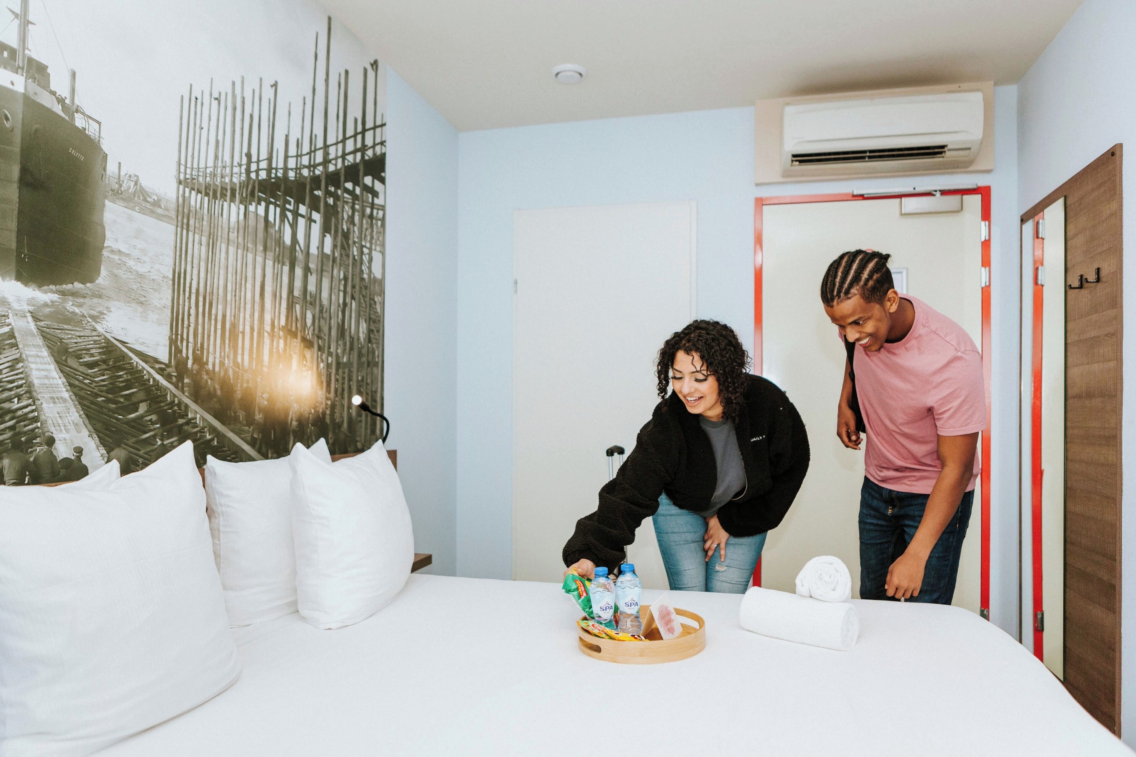Habitación de hotel con cama, almohadas, un mural en la pared y una mesita de madera con aperitivos y agua embotellada.