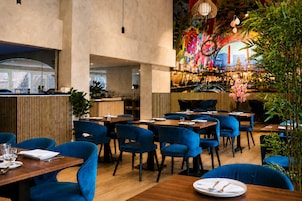 Un interior de restaurante con sillas tapizadas en azul, mesas de madera y un mural colorido en la pared.