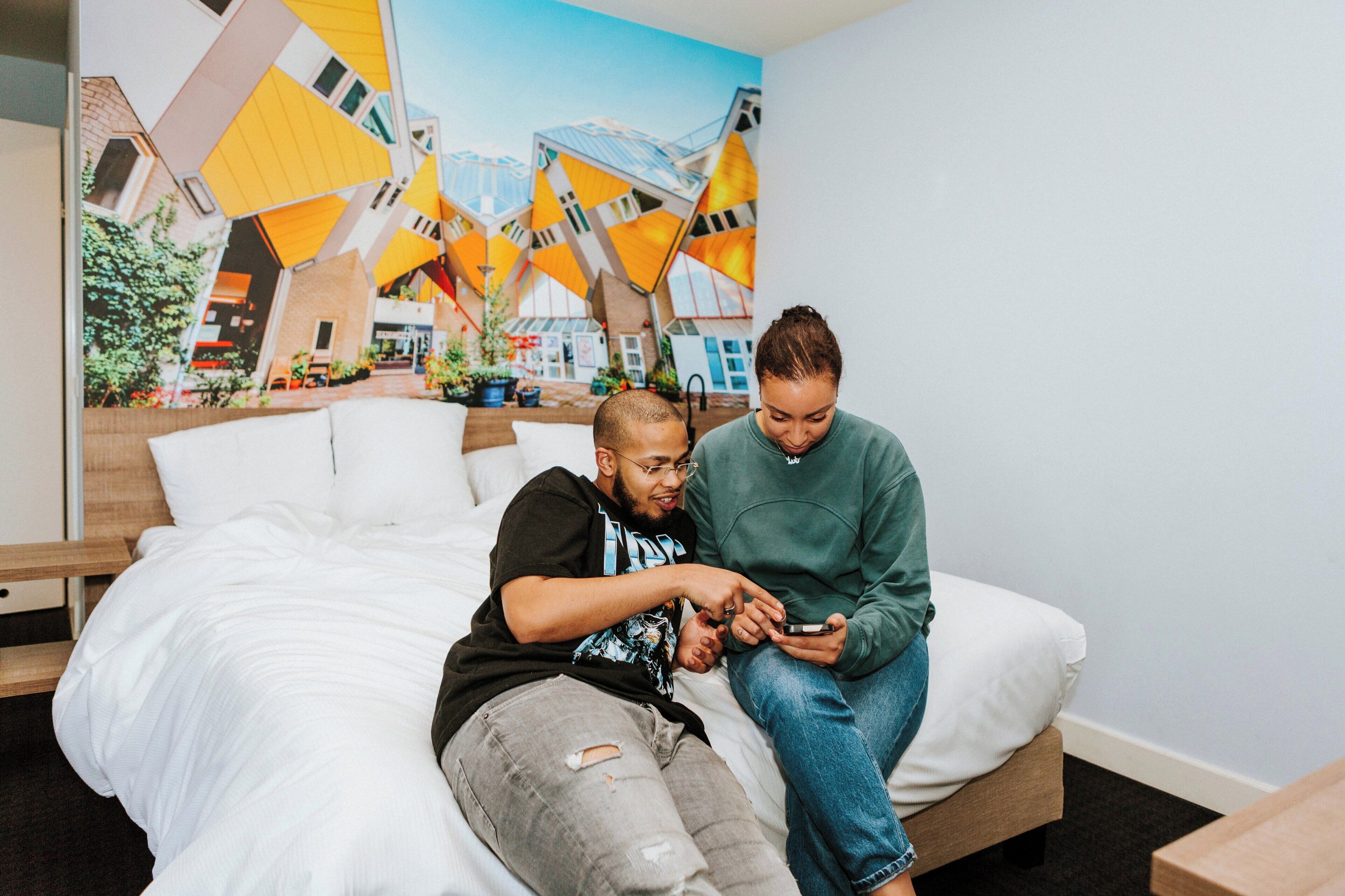 A bed with a white comforter and pillows, a wooden headboard, and a wall mural of a colorful building.