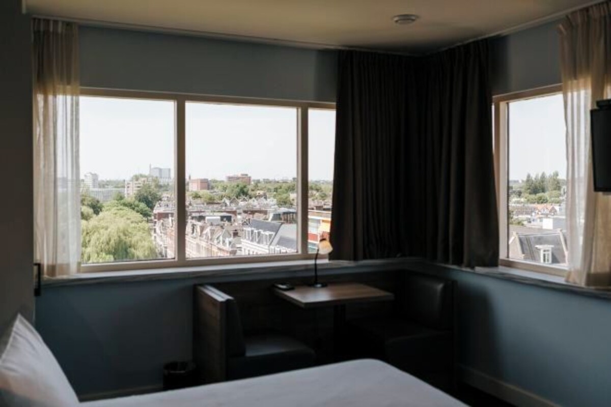 Habitación de hotel con un ventanal grande que ofrece vistas a la ciudad, una cama, un escritorio y una silla.