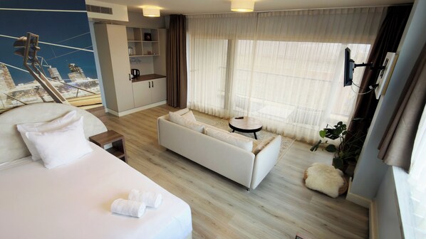 A hotel room with a bed, a sofa, a small table, a coffee machine, and a large window with curtains.