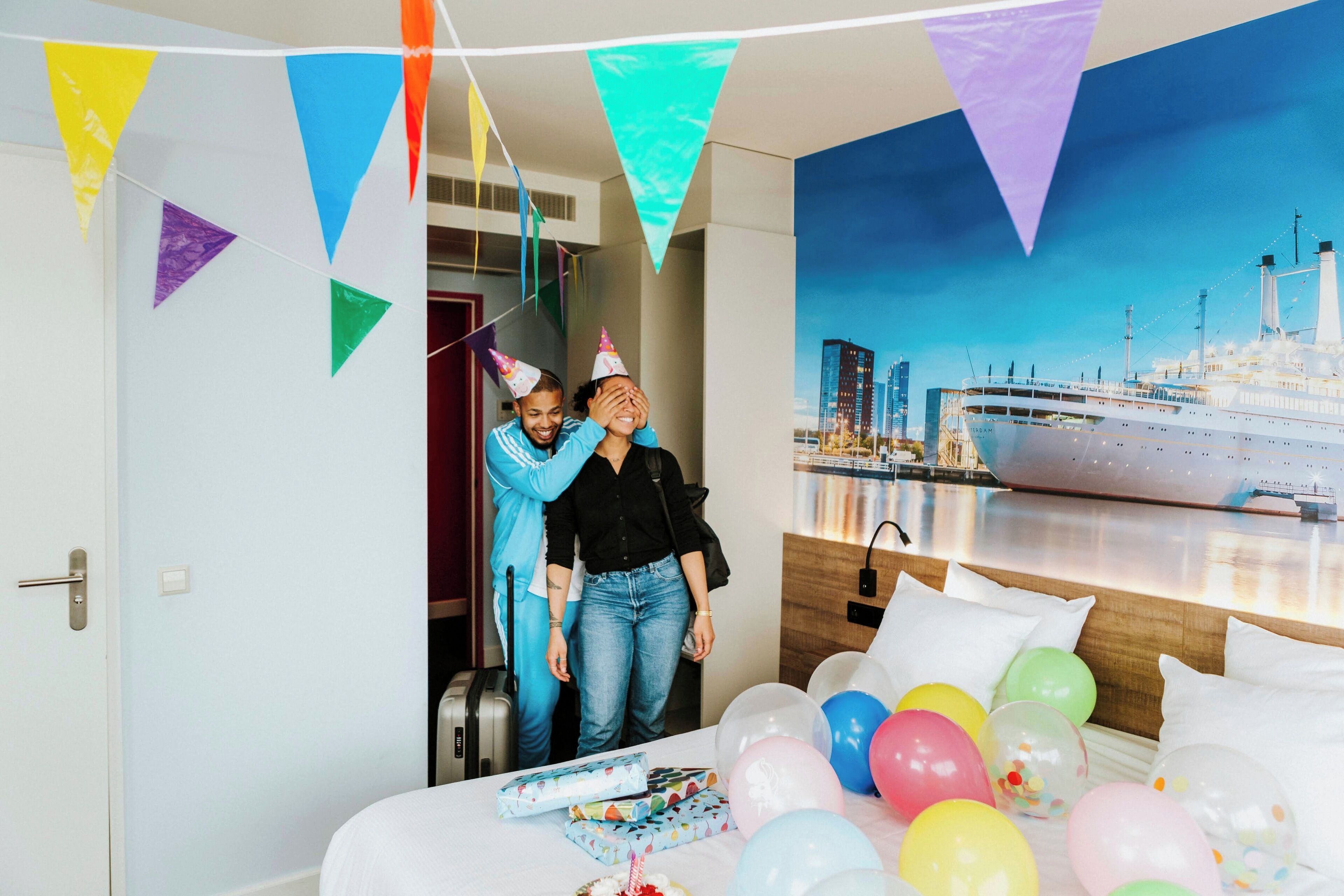 Ein Zimmer mit einem Bett, einem Tisch mit Luftballons und einem Stadtbild-Wandgemälde.