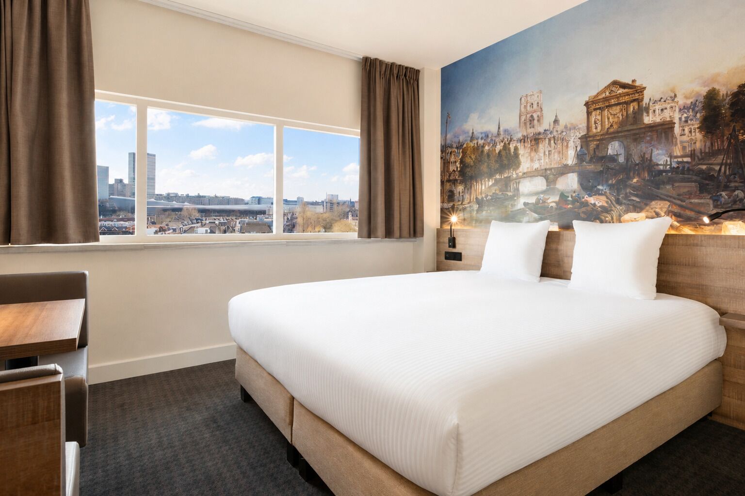 Ett hotellrum med en stor säng, utsikt över staden och en målning av en historisk byggnad.