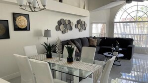 Een moderne woonkamer met een glazen eettafel, witte stoelen, een zwarte bank en een plafondventilator.