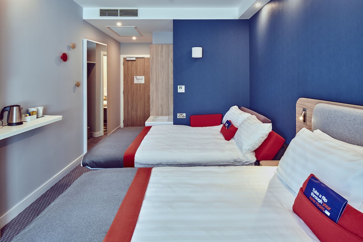 Une chambre d’hôtel avec deux lits, une décoration aux couleurs rouge et blanche, une petite kitchenette et une porte en bois.