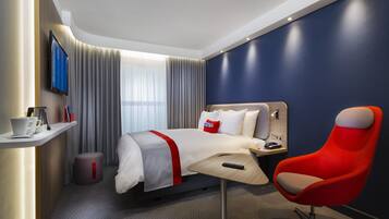A modern hotel room with a bed, a red armchair, a TV, and a small table with a remote control.