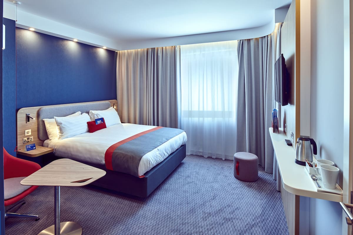 Une chambre d’hôtel moderne avec un lit bien fait, un bureau doté d’un téléviseur à écran plat, une chaise et un petit tabouret.