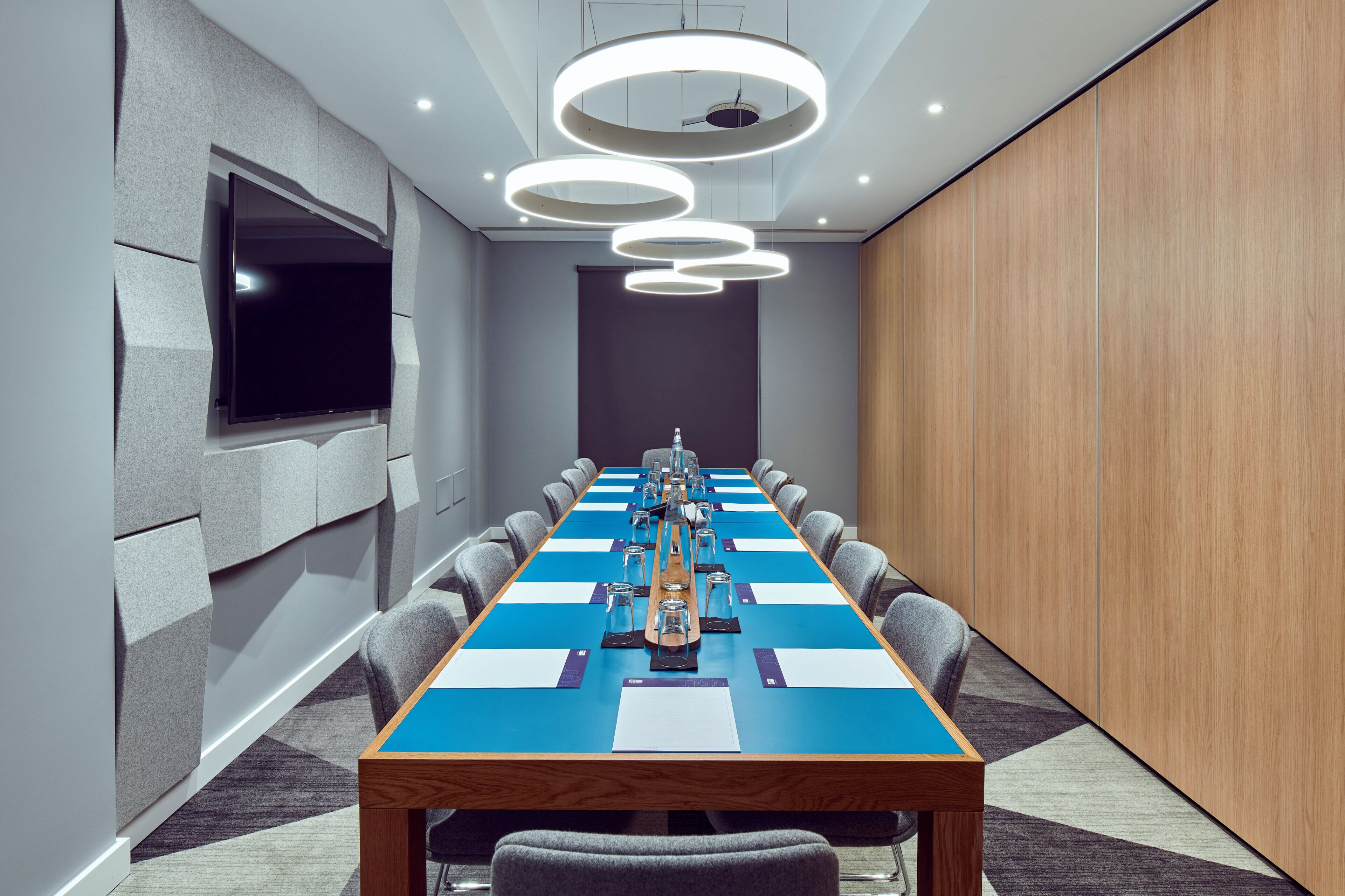 A conference room with a long table, chairs, a flat-screen TV, and modern lighting fixtures.