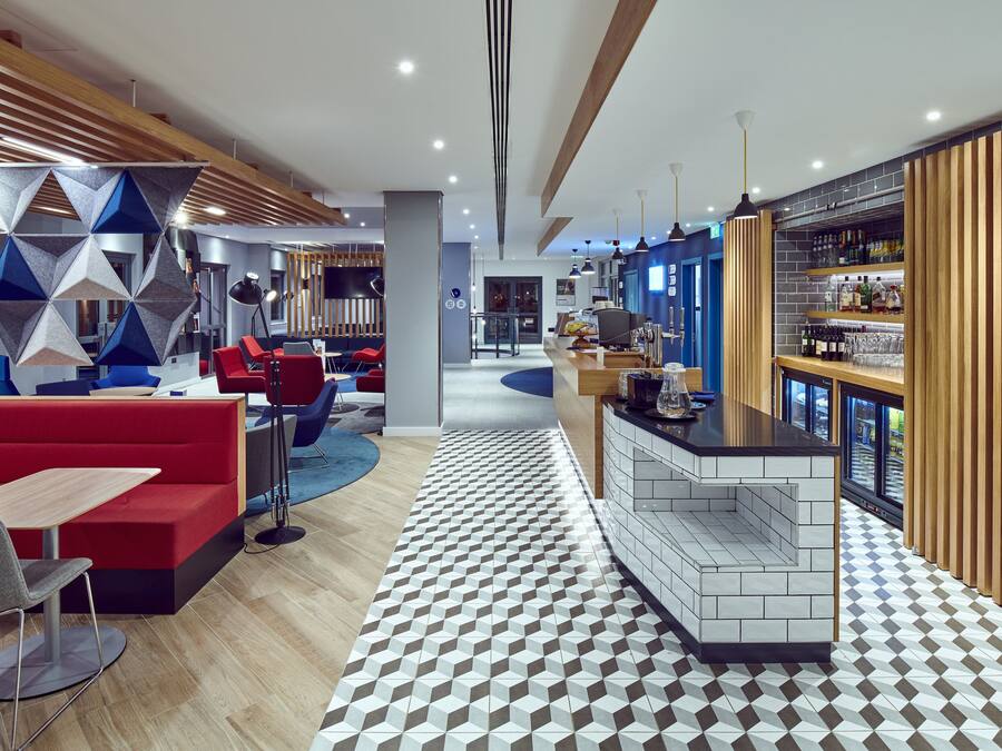 A modern hotel lobby with a geometric patterned floor, a bar area with a tiled backsplash, and a seating area with red sofas and a blue circular rug.