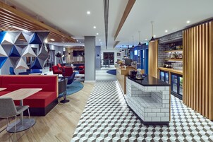 Un moderno vestíbulo de hotel con piso de diseño geométrico, una zona de bar con salpicadero de azulejos y un área de estar con sofás rojos y alfombra azul circular.