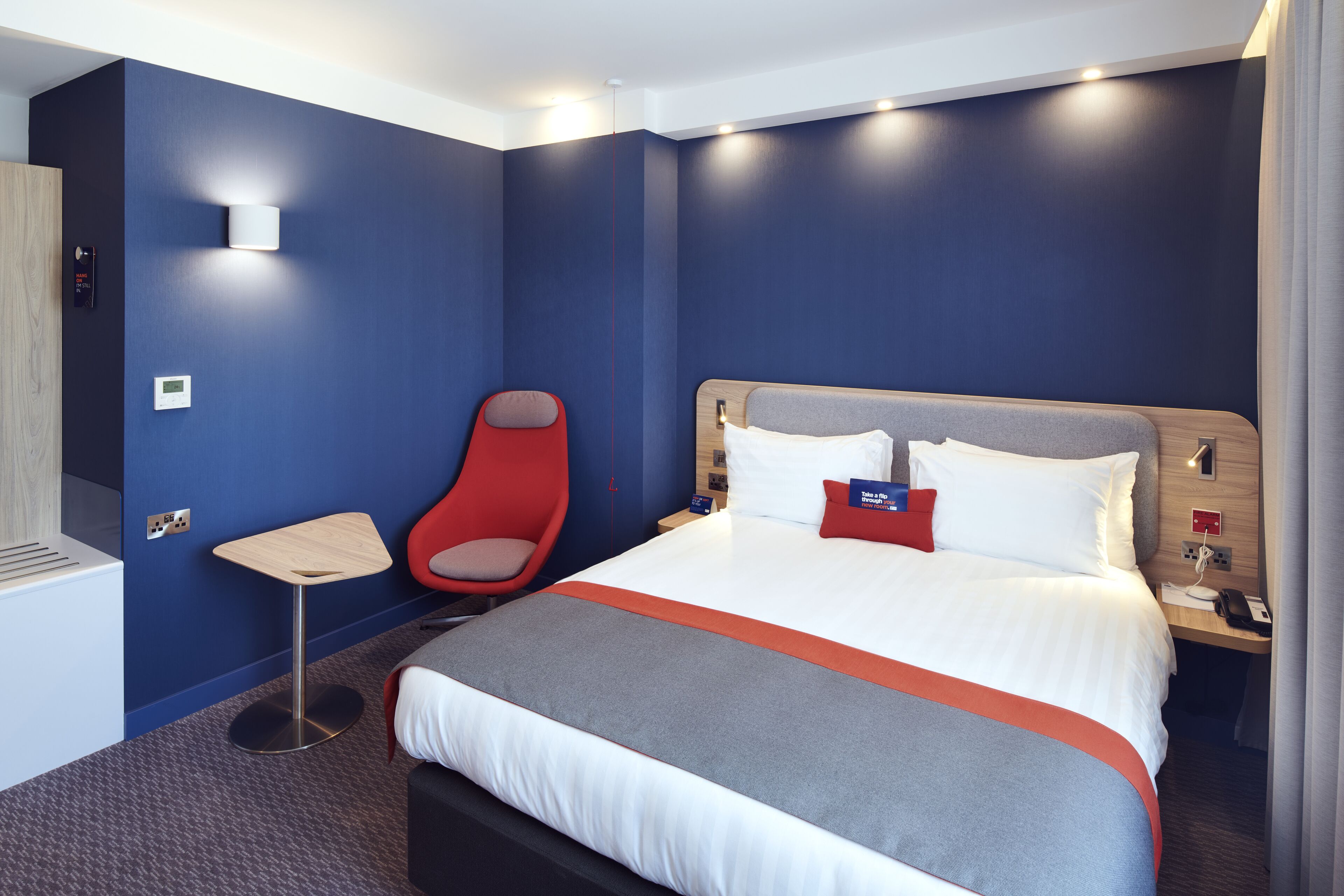 Ein modernes Hotelzimmer mit einem großen Bett, einem roten Sessel, einem kleinen Tisch und einem Nachttisch aus Holz.