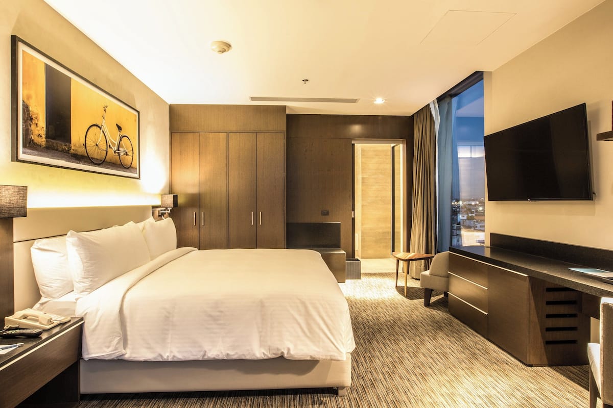 A modern hotel room with a large bed, a desk, a TV, and a painting on the wall.